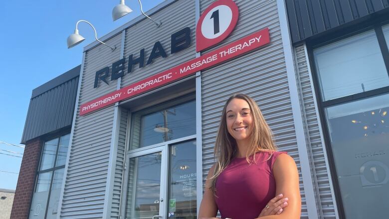 a woman with her arms crossed, smiling standing in front of a business with a sign that reads Rehab 1, physiotherapy, chiropractic, massage therapy