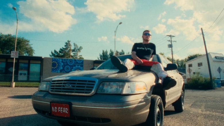 A man sits on a car in a parking lot on a sunny day.