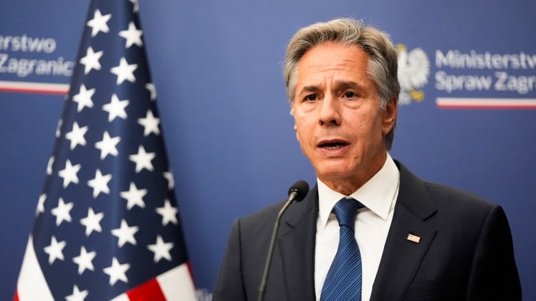 A gray-haired man in a dark suit and blue tie speaks into a microphone. An American flag is behind him.