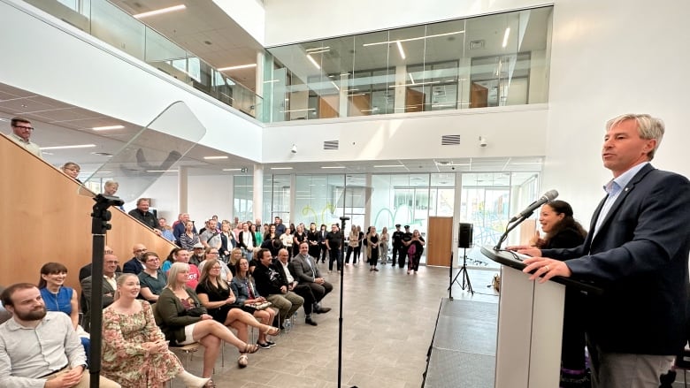 A man speaks at a podium on the right in front of a small crowd in a large open room with large windows and natural light.