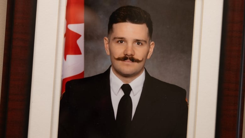 A portrait of a young firefighter wearing his dress uniform, standing in front of a Canadian flag. 