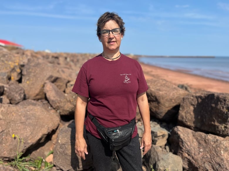 A woman with short reddish hair and glasses wearing a burgundy t-shirt standing on a beach.