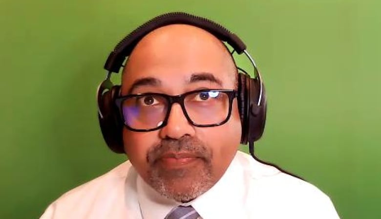 A man wearing a white shirt and tie sits in front of a green screen.