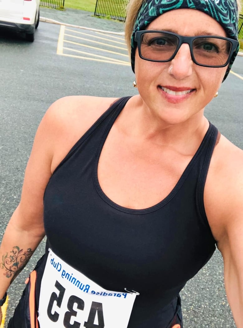 A woman in a black tank top and sunglasses smiles for the camera. She is wearing a runners number bib and a green bandana.