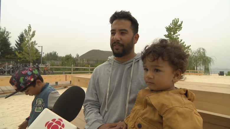 A man and two young children sit on a bench by a beach on a foggy grey day, speaking to a reporter's microphone