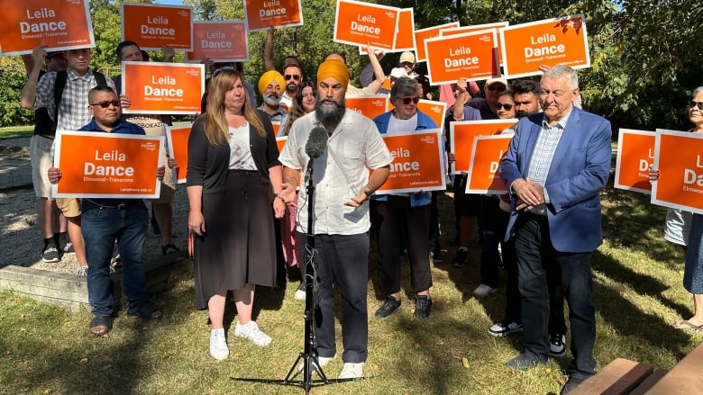 Two people stand close to a microphone in front of a crowd of people holding orange coloured signs. 