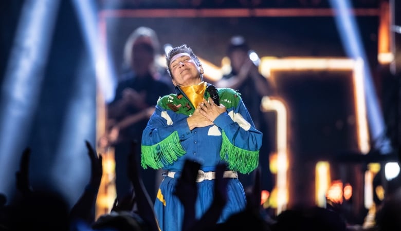 A singer in a blue dress with green fringe holds her hands to her chest in a loving motion.