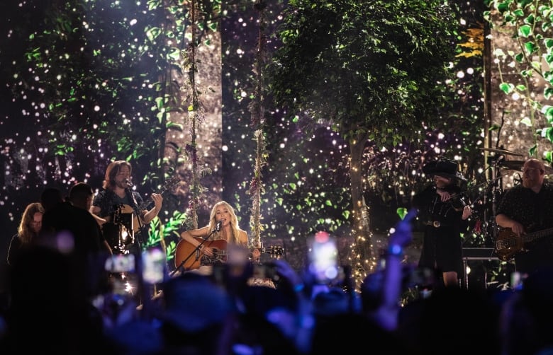 A blonde singer sits on a stool and plays guitar as lights twinle in the background.