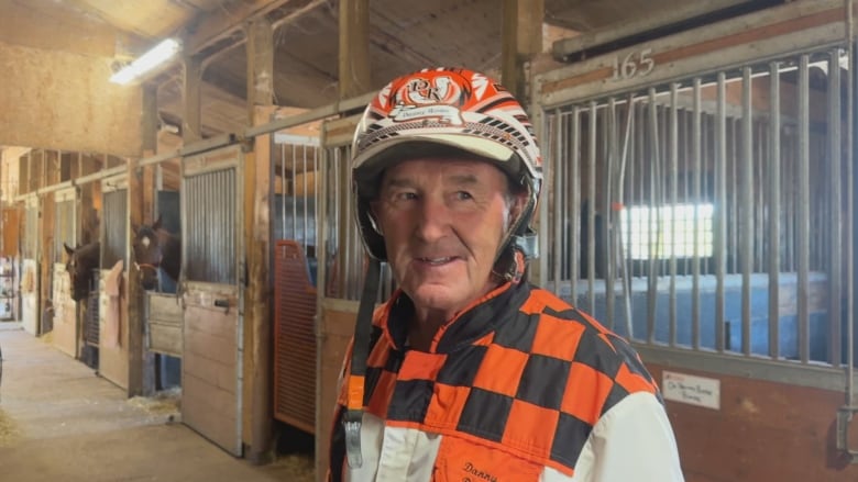 A harness racer in helmet and an orange and black riding top looks at the camera.