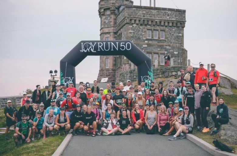 A large group of runners gather in front of a race starting line for a group photo. There is a large black archway behind them that reads Joy Run 50.