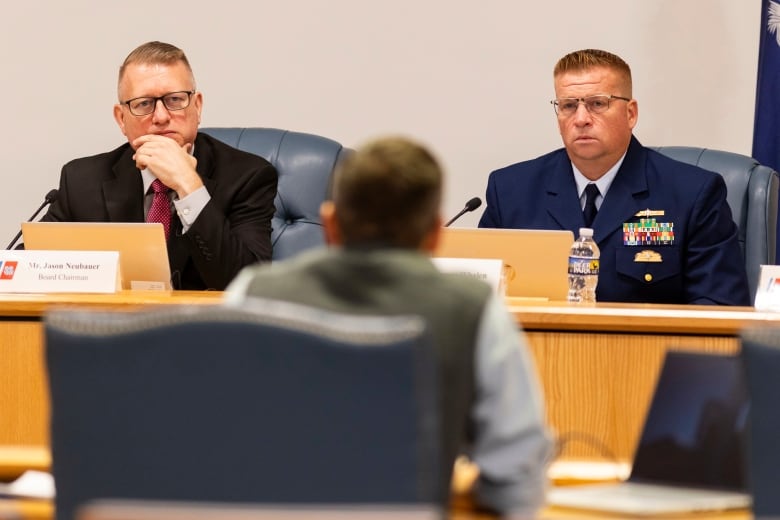 A seated man in a dark suit and a man in a blue uniform listen to a man speak.