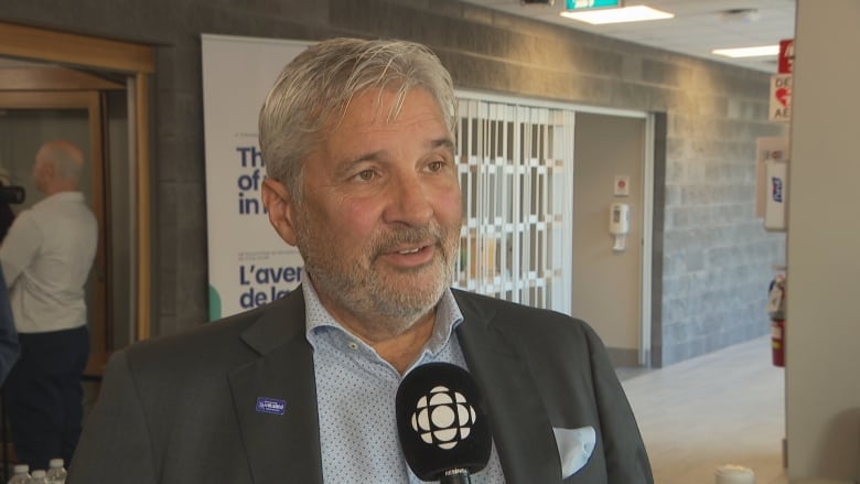 A man with grey hair in a grey suit jacket and blue button up shirt speaks into a microphone.