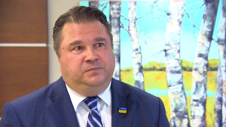 A man in a suit does in interview in front of a backdrop of trees.