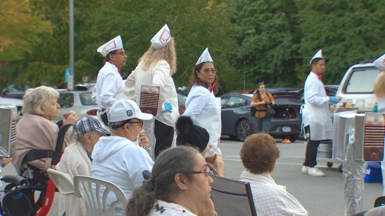 A number of people are seen wearing white costumes with white hats, in the style of old cinema attendants. One of them is pictured at a table serving food.