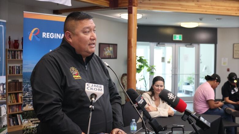 Man wearing a black jacket stands in front of microphones at a lecturn. People sit in the background