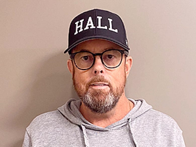 A man in a grey hoodie with black glasses wears a blue ballcap that says 