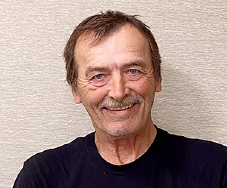 A man with short dark hair and a grey moustache wears a dark shirt and smiles.