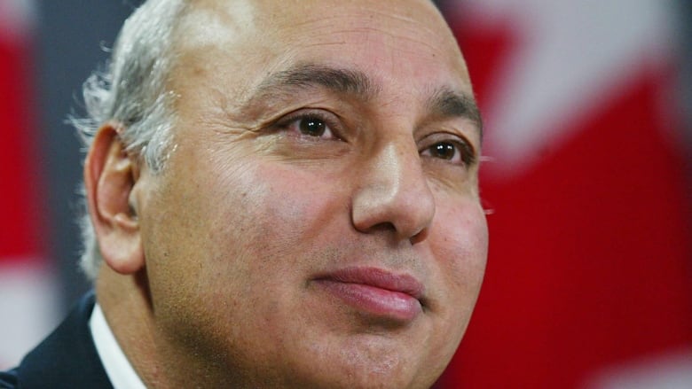 A man with white hair and a round face with a hint of a smile poses in front of a Canadian flag in this head-and-shoulders shot.