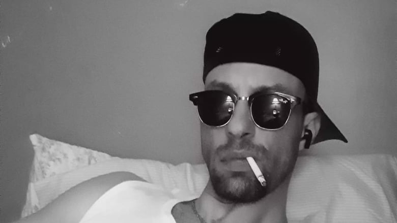 Man in sunglasses, ballcap on backwards and a cigarette hanging out of his mouth, reclining on a pillow. 