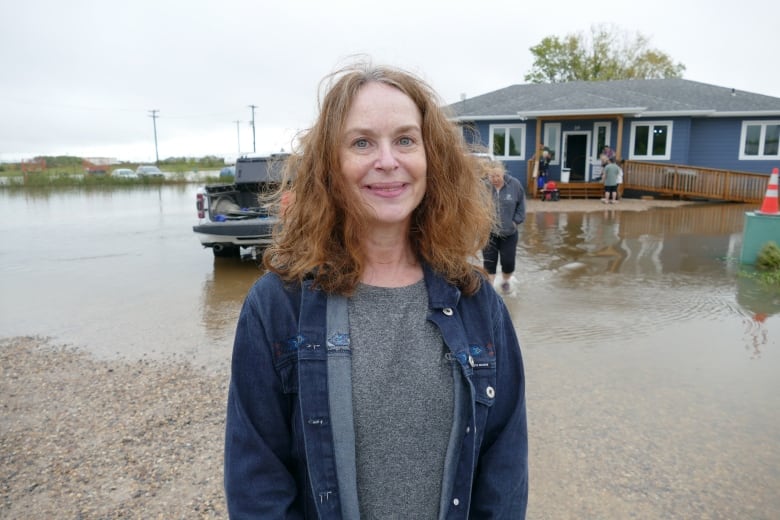 Woman looks at the camera with a smile, behind her a building is surrounded by water after overland flooding. 