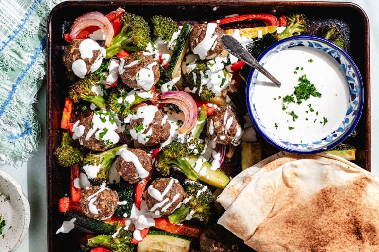 Overhead shot of a sheet pan with meatballs, roasted vegetables, pitas and a small bowl of yogurt tahini sauce.