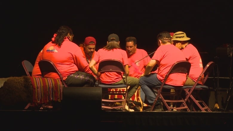 A group of people wearing orange shirts gather around a drum while performing