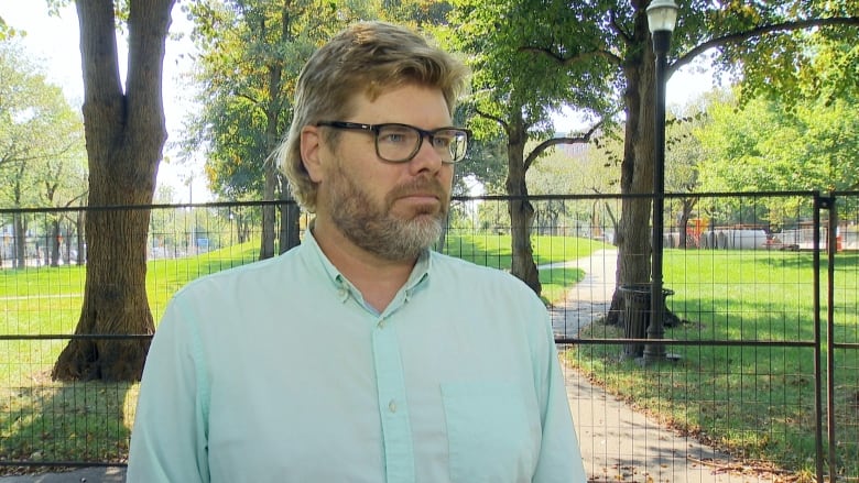 A white man with blonde hair, glasses and a beard wears a light green button-up shirt. He stands in front of black fencing with a grassy park behind it