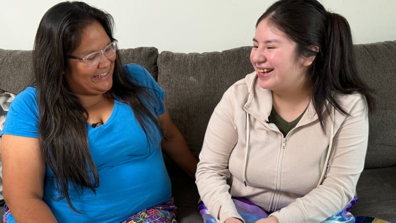 Two women in ribbon skirts sit side by side on a couch.