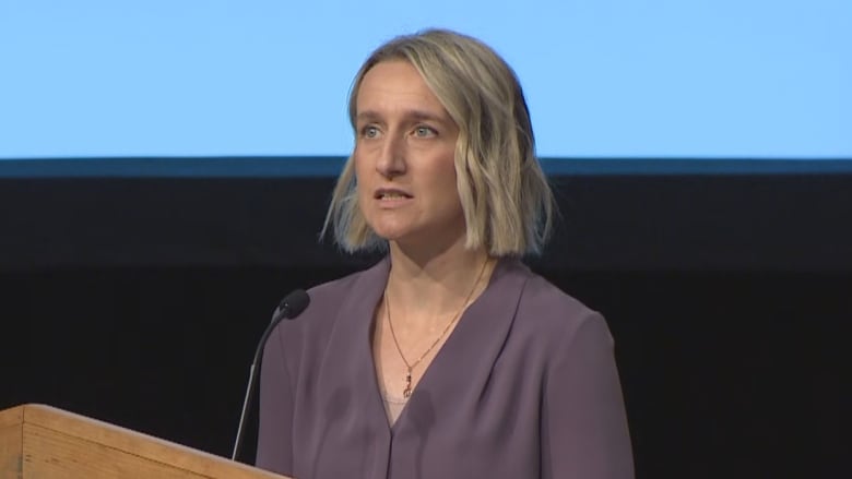 A woman with blond hair in a purple shirt stands at a podium with a blue screen behind her.