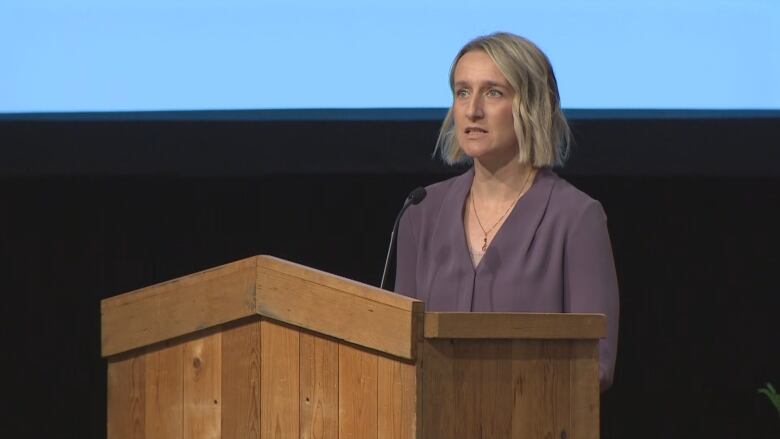 A woman with blond hair in a purple shirt stands at a podium with a blue screen behind her.