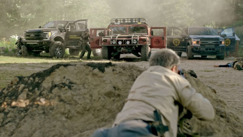 A man in a beige jacket with his back to the camera crouches in a hole and points a weapon at armed people emerging from three huge trucks.