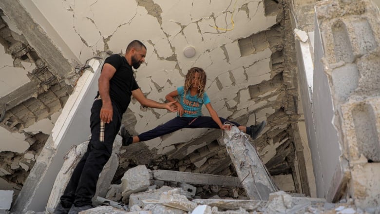 A boy does the splits on a destroyed building with his father standing next to him.