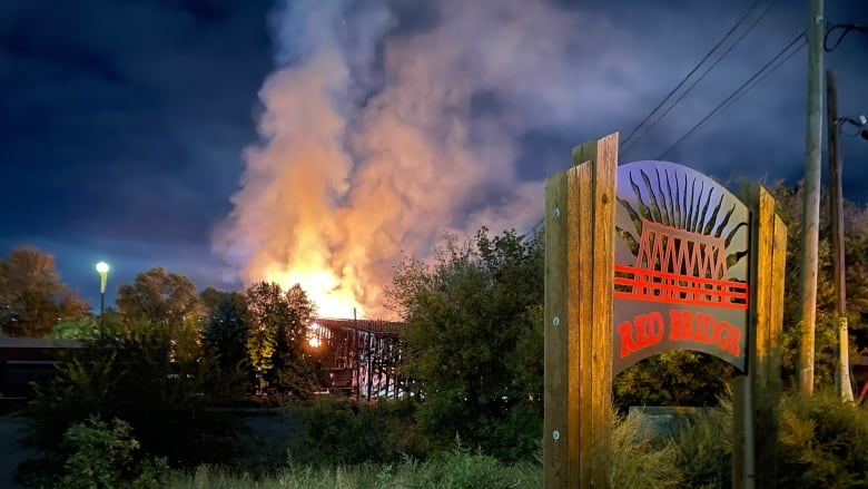 A sign for the Red Bridge in the foreground right, with the structure in flames in the distance.