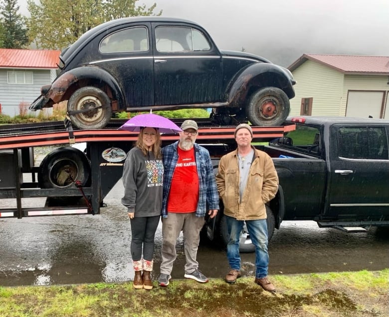 A women and two men standing in front of a pickup truck with a trailer carrying a vintage Volkswagen Beetle. 