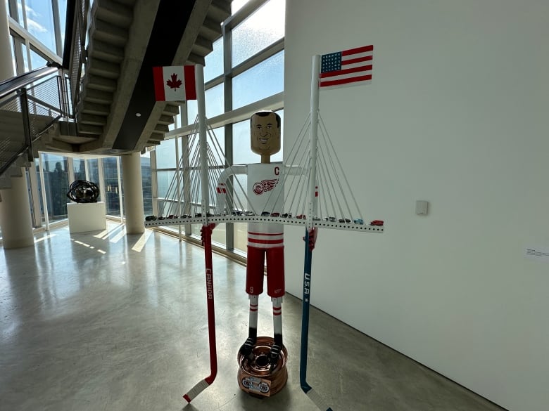 A metal figure looking like hockey player Gordie Howe holds on to two sticks that are holding up a bridge with mini cars on it.