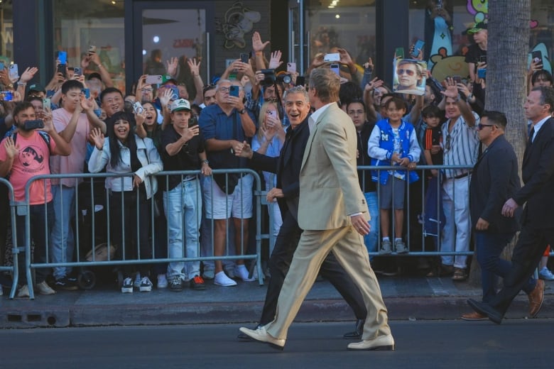 Two smiling men in suits walk on a street. Behind them is a large crowd of people cheering. 