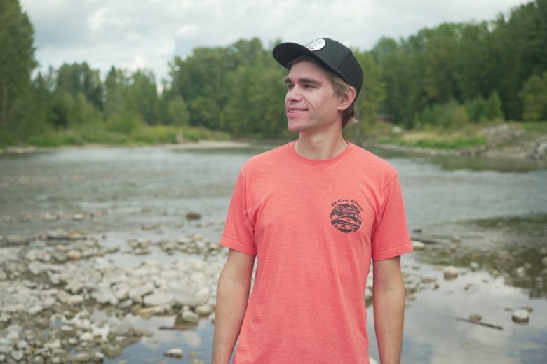 A young man in a red T-shirt and a black baseball cap stands on the rocky shore of a river. 