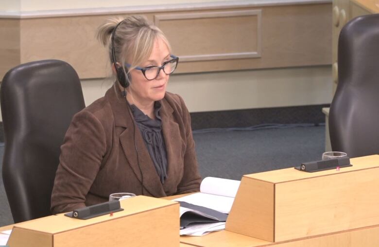 A blonde woman wearing a brown corduroy jacket over a frilly black shirt, headphones and glasses sitting at a curved table