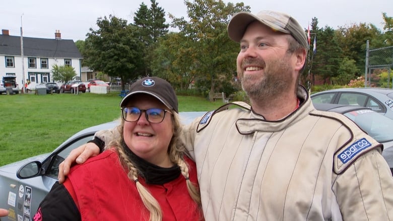 Man stands with arm around woman smiling. Race car is behind them.