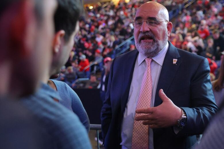 Late-middle-aged bald man in a suit, chatting with people on the floor of a dimly lit convention arena