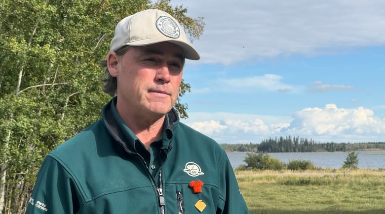 A white man is standing outside in a nature park; white puffy clouds sit behind him in the blue sky. The man is wearing a beige cap and a dark green jacket.