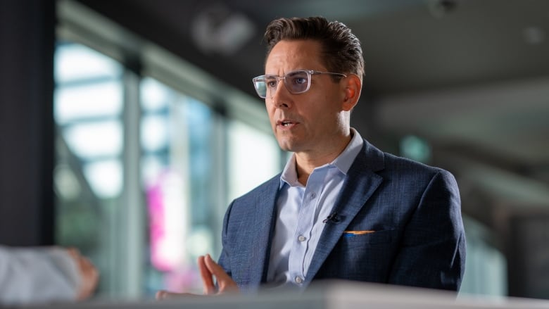 A man in a blue suit jacket with dark hair and glasses looks across a desk