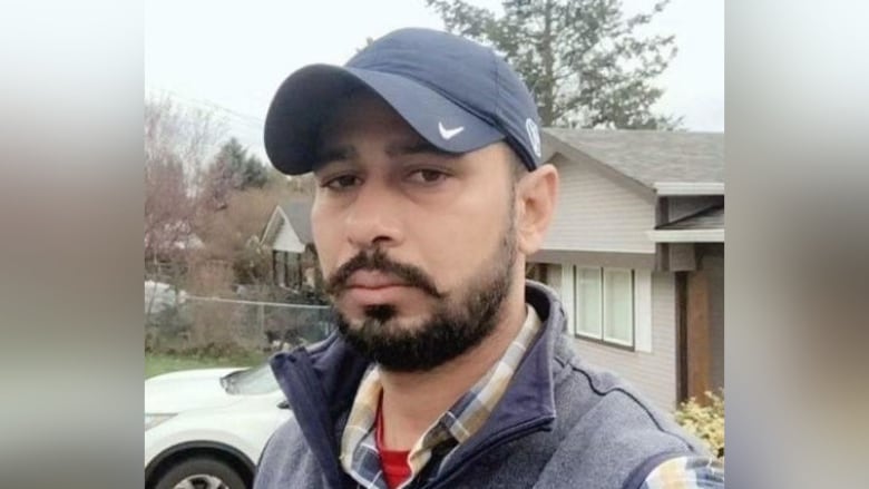 A man with beard wears a hat with a checkmark on the brim and a plaid shirt and vest. 