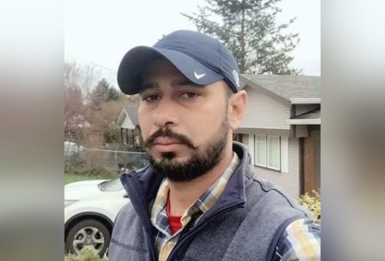 A man with beard wears a hat with a checkmark on the brim and a plaid shirt and vest. 