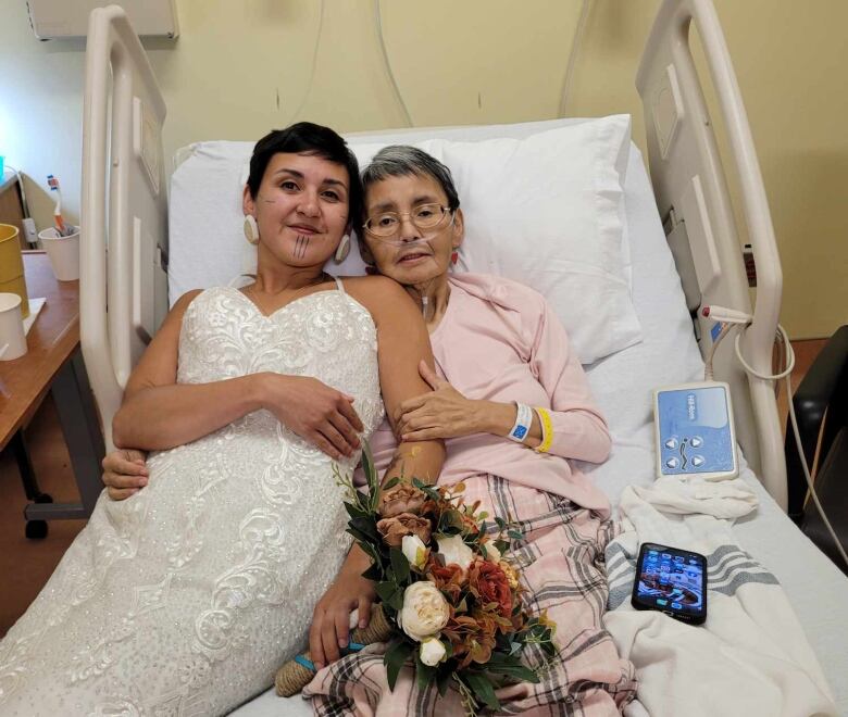 Woman with short dark hair in a wedding dress reclining in a bed with an older woman in pink pajamas.