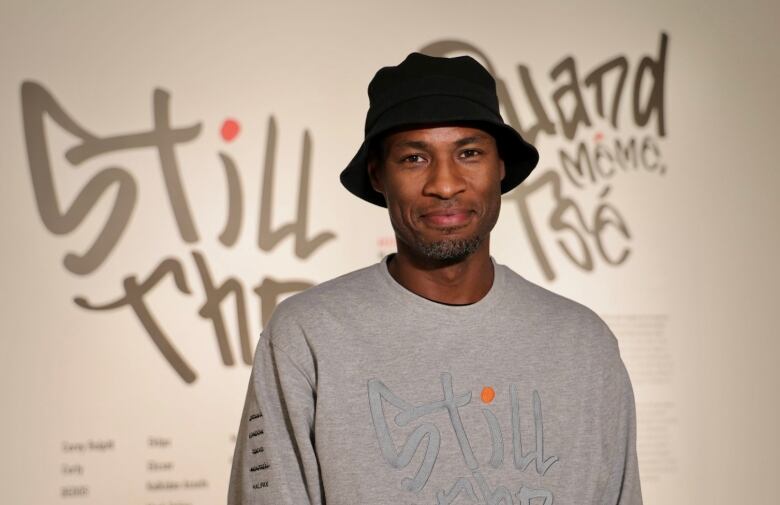 A man with a hat stands in front of a wall with graffiti-style writing.