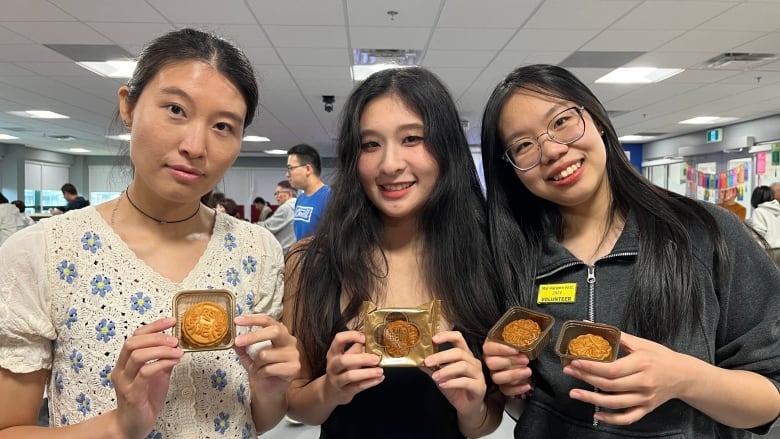 Three women with black hair stand next to each other smiling showing the mooncakes in their hands. 