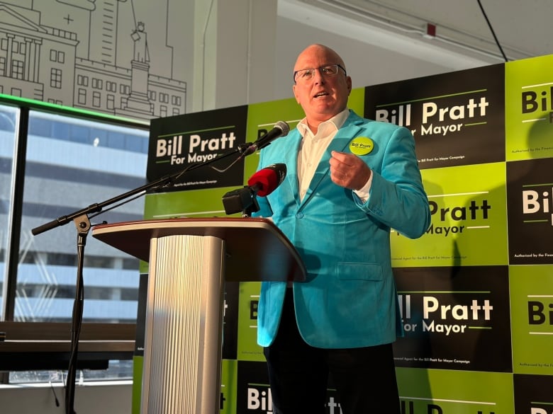 A man stands at a podium. He is wearing a white button up shirt and a blue blazer. Behind the man is a wall with signs reading 