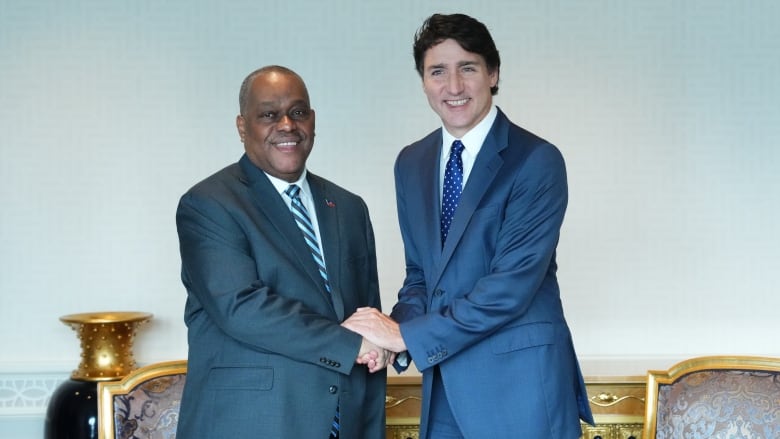 Prime Minister Justin Trudeau and Haiti Prime Minister Garry Conille shake hands. Both men are wearing suits and are shown standing in front of two arm chairs. 