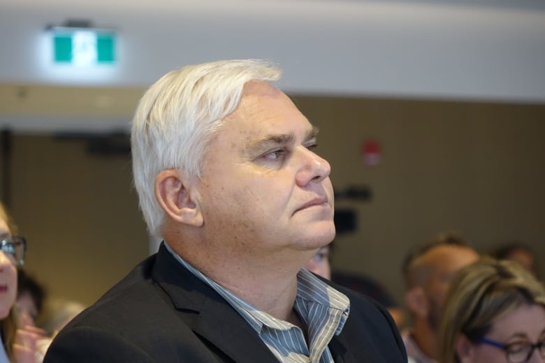 A man wearing a black blazers listens intently in a crowd.